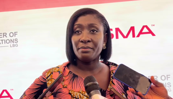 African woman in a colorful floral dress speaks at a press conference, microphones in front, a person holds a phone toward her.