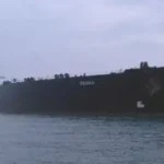 Large black cargo ship moored in calm water with an overcast sky and a second vessel visible in the background on the left.