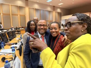 Four colleagues take a selfie at a conference table in a meeting room; a smiling man in a suit, two women, and a person in a bright yellow jacket taking the photo.