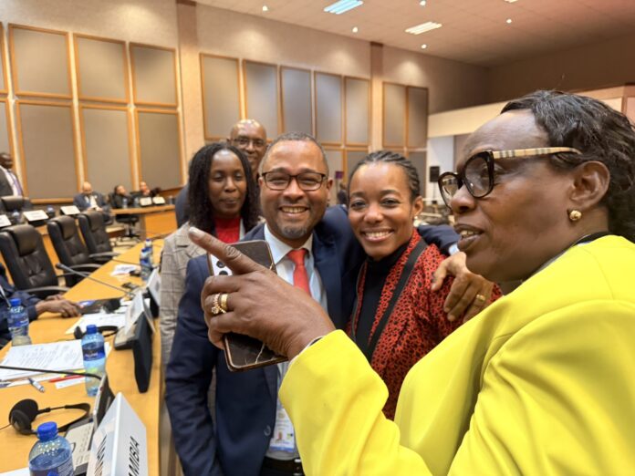 Four colleagues take a selfie at a conference table in a meeting room; a smiling man in a suit, two women, and a person in a bright yellow jacket taking the photo.