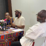 Man speaking at a press conference with multiple microphones on a colorful patterned tablecloth; two other men listen attentively nearby.