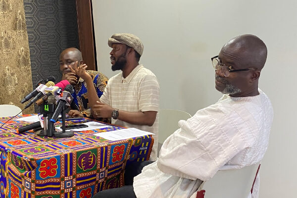 Man speaking at a press conference with multiple microphones on a colorful patterned tablecloth; two other men listen attentively nearby.