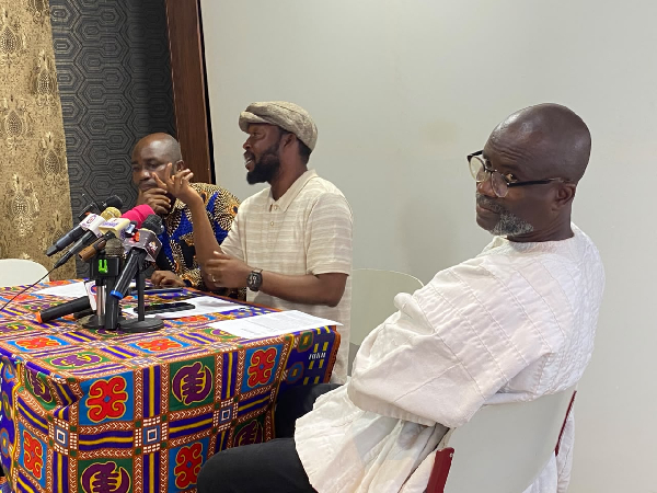 Man speaking at a press conference with multiple microphones on a colorful patterned tablecloth; two other men listen attentively nearby.