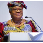 African woman in colorful traditional dress speaking at a podium with a microphone, delivering a speech at a formal event.