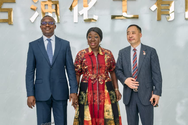 Three professionals posing for a photo in a corporate lobby, two men in suits flanking a woman in a colorful patterned dress, gold characters on the wall behind them.