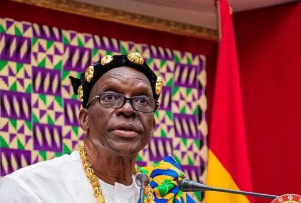 Man in white traditional outfit with gold beads and headpiece speaks at a podium with a colorful geometric backdrop.