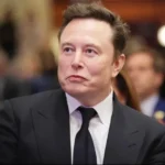 Man in a black suit and tie at a formal event, looking to the side with a neutral expression.