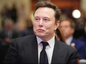 Man in a black suit and tie at a formal event, looking to the side with a neutral expression.