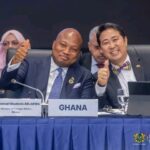 Two men in suits at a formal conference smile and thumbs up; a sign reading GHANA and a Ministry of Foreign Affairs logo are visible on the table.