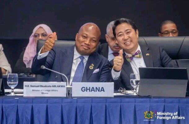 Two men in suits at a formal conference smile and thumbs up; a sign reading GHANA and a Ministry of Foreign Affairs logo are visible on the table.