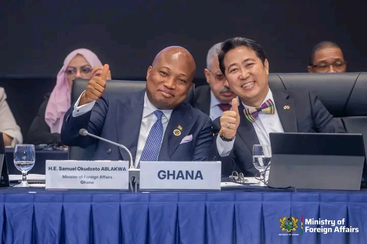 Two men in suits at a formal conference smile and thumbs up; a sign reading GHANA and a Ministry of Foreign Affairs logo are visible on the table.