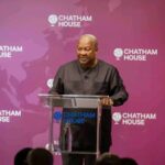 Man in a black traditional shirt speaking at a clear podium during a Chatham House event, with a purple backdrop and logos behind him.