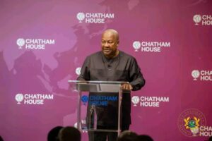 Man in a black traditional shirt speaking at a clear podium during a Chatham House event, with a purple backdrop and logos behind him.