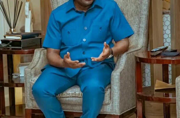 Man in a blue short-sleeve shirt and pants sits in a beige armchair, gesturing with his hands while talking in a living room setting.