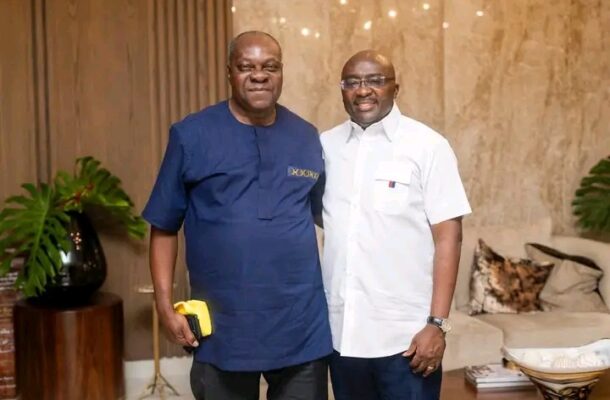 Two men pose for a photo indoors; one in a blue traditional shirt holds a yellow item, the other in a white short-sleeve shirt, with plants and a sofa in the background.