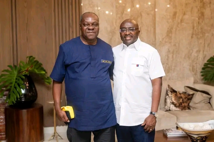 Two men pose for a photo indoors; one in a blue traditional shirt holds a yellow item, the other in a white short-sleeve shirt, with plants and a sofa in the background.