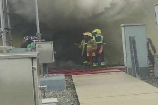 Firefighters in yellow helmets and turnout gear stand at a smoke-filled doorway, inspecting a red fire hose as thick dark smoke billows from a building.