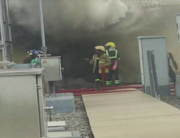 Firefighters in yellow helmets and turnout gear stand at a smoke-filled doorway, inspecting a red fire hose as thick dark smoke billows from a building.