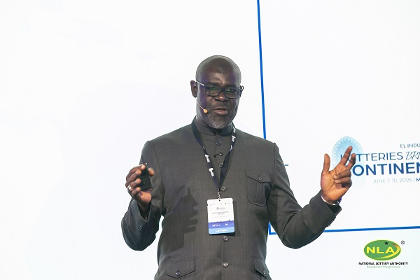 African man in a dark suit giving a presentation on stage, wearing a headset mic and conference badge.