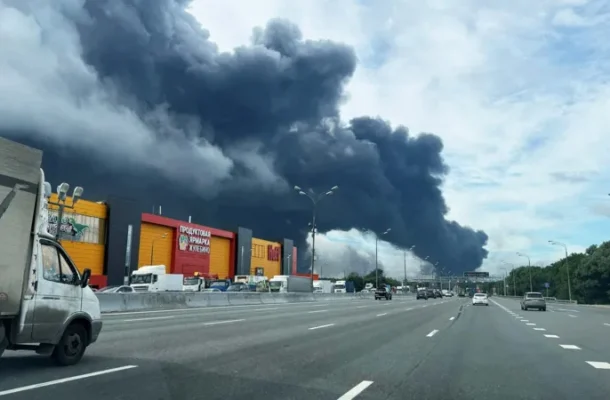 Thick black smoke rises from a fire behind a row of shops along a multi-lane highway; vehicles drive by on the road far from the blaze.