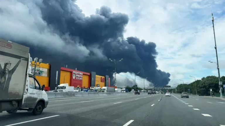 Thick black smoke rises from a fire behind a row of shops along a multi-lane highway; vehicles drive by on the road far from the blaze.