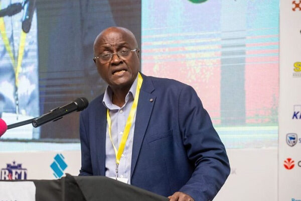 38017276 | Ghanaguardian.com Black man in a navy suit delivers a speech at a conference from a podium with a microphone, wearing a yellow lanyard; backdrop shows logos and screens.