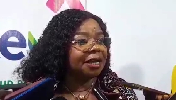 Woman with curly hair and glasses speaks at a press event with multiple microphones in front of her background logos.