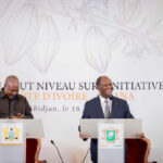 Two men in suits speak at podiums during a formal event, with an Ivory Coast cacao initiative banner and crest emblems behind them.