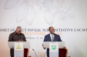 Two men in suits speak at podiums during a formal event, with an Ivory Coast cacao initiative banner and crest emblems behind them.