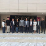 Twelve professionals in business attire pose for a group photo on marble steps outside a building with a black plaque featuring gold calligraphy.