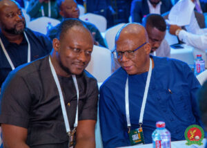Two men wearing lanyards sit side by side at a conference, one in a black shirt and the other in blue, conversing.
