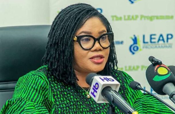 African woman with black glasses speaks at a press conference, green patterned outfit, microphones in front, LEAP logo backdrop.