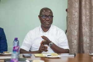 Man in a white short-sleeve shirt and glasses sits at a table in a meeting, hands clasped, with a water bottle and papers in front of him.