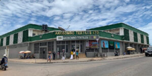 Storefront of Akosombo Textiles Ltd, a green, single-story shop on a busy street with pedestrians and motorcycles and a bright blue sky behind it.