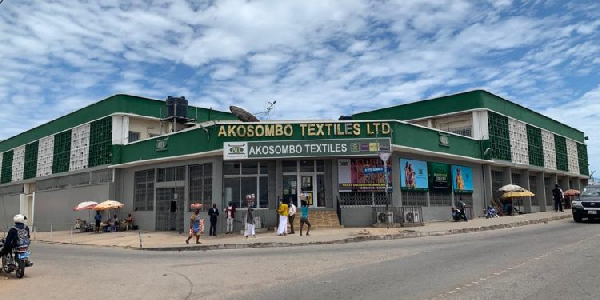 55746949 | Ghanaguardian.com Storefront of Akosombo Textiles Ltd, a green, single-story shop on a busy street with pedestrians and motorcycles and a bright blue sky behind it.