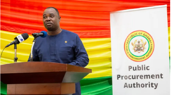 56096933 | Ghanaguardian.com Man in a blue outfit speaking at a wooden podium, with a red-yellow-green Ghana flag backdrop and a banner reading Public Procurement Authority.
