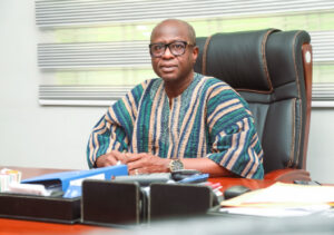 Man in a colorful striped traditional shirt sits at a wooden desk in an office, facing the camera.
