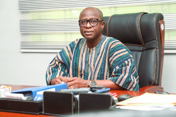 Man in a colorful striped traditional shirt sits at a wooden desk in an office, facing the camera.