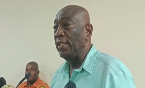 Man in a teal shirt speaks into a microphone at a press conference, with another man in orange in the background.