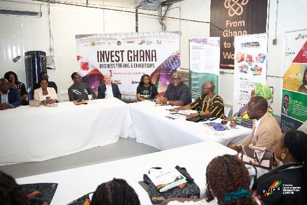 Group of diverse professionals seated around a white-clothed conference table at an Invest Ghana business event, with banners and posters in the background.