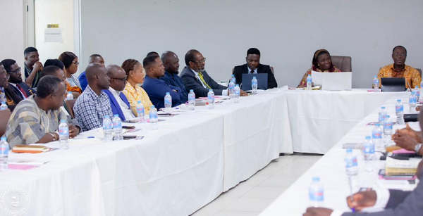 A group of professionals seated around U-shaped white-clothed conference tables in a meeting room, with water bottles and notebooks on the table.