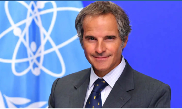 Smiling middle-aged man in a dark suit and blue tie stands in front of a blue backdrop with white orbital lines behind him.