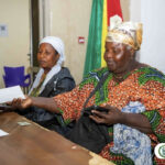 Two women sit at a table with papers; the woman in a colorful patterned dress extends her hand toward a document while the other watches, with the ACCRA Metropolitan Assembly logo in the corner.