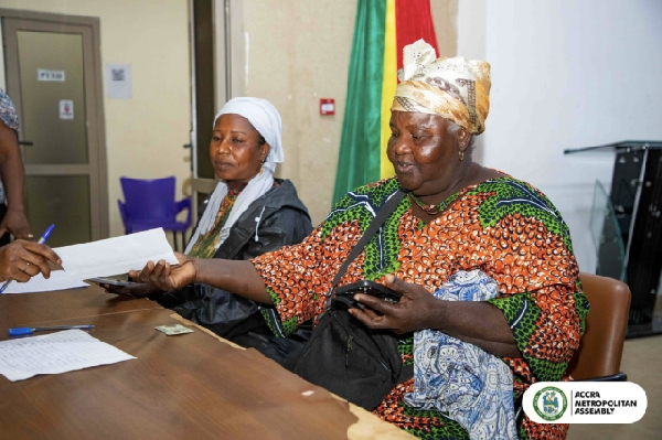 Two women sit at a table with papers; the woman in a colorful patterned dress extends her hand toward a document while the other watches, with the ACCRA Metropolitan Assembly logo in the corner.