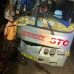 Front of a gray Intercity bus with a severely cracked windshield and scattered debris after a collision, nighttime scene.