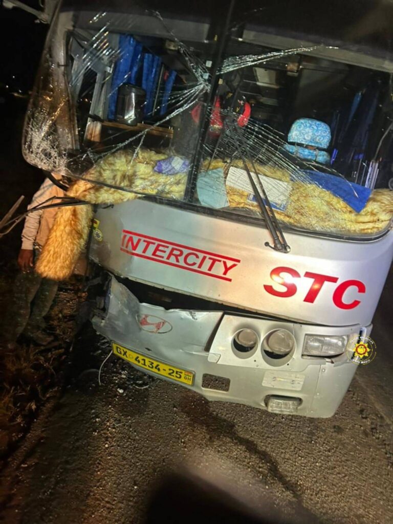 Front of a gray Intercity bus with a severely cracked windshield and scattered debris after a collision, nighttime scene.