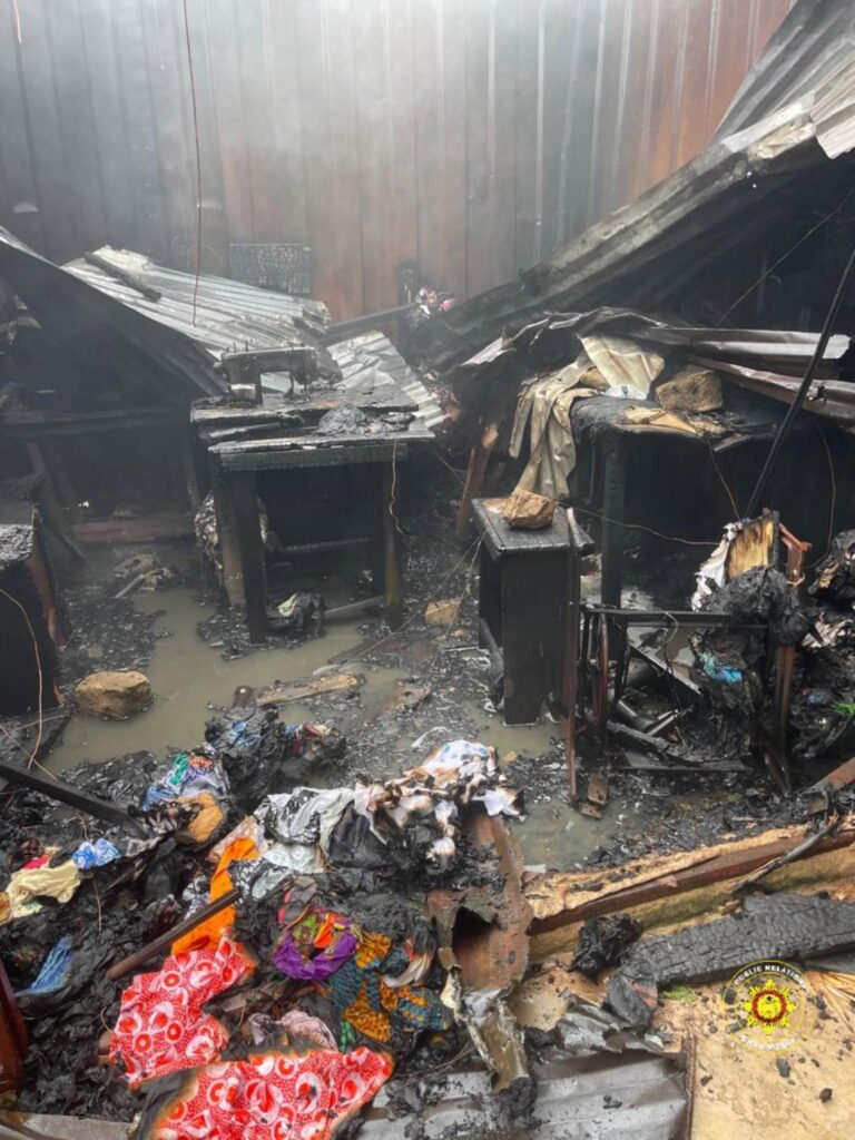 Fire-damaged room with a collapsed roof, charred furniture, and standing muddy water; scattered burnt debris and colorful fabric in the foreground; fire department logo visible bottom right.