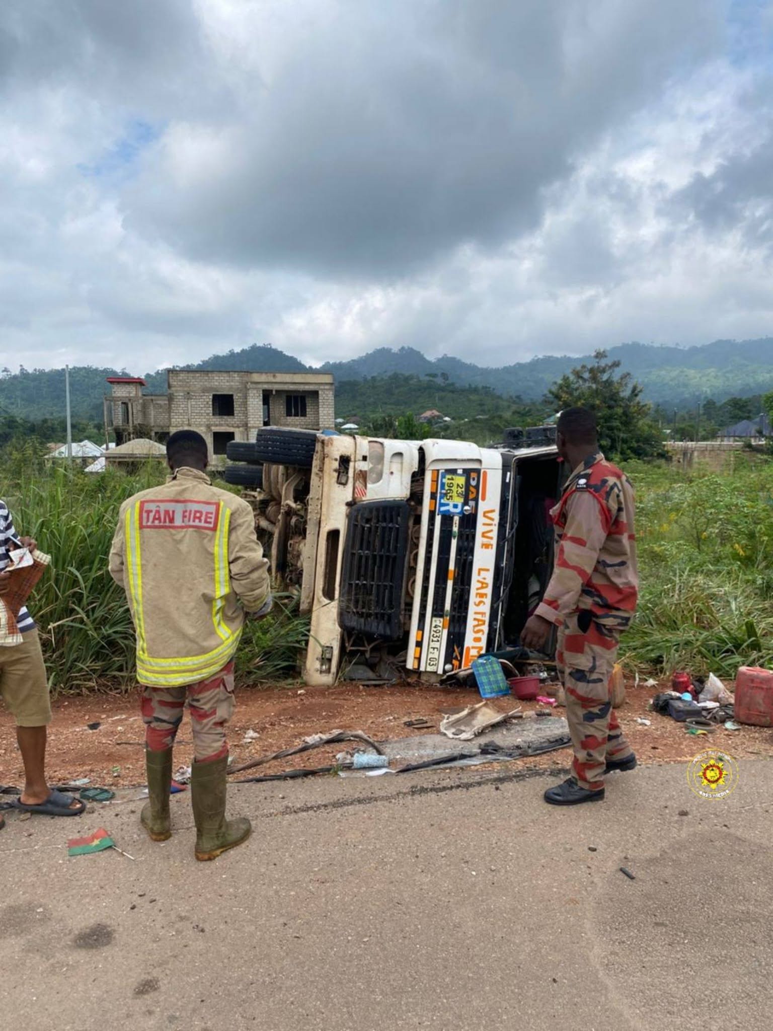 721430436_1018456804026606_9127948363394490276_n | Ghanaguardian.com Firefighters in tan uniforms inspect an overturned white truck on a rural road; debris lies nearby, with a half-built brick building and green hills in the background.