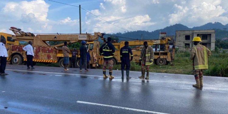People in work clothes and firefighters stand on a wet highway beside a large yellow crane truck; hills and a building under construction are in the background.