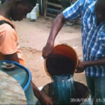 Two men pour a dark liquid from an orange bucket into a blue barrel in an outdoor workshop area.
